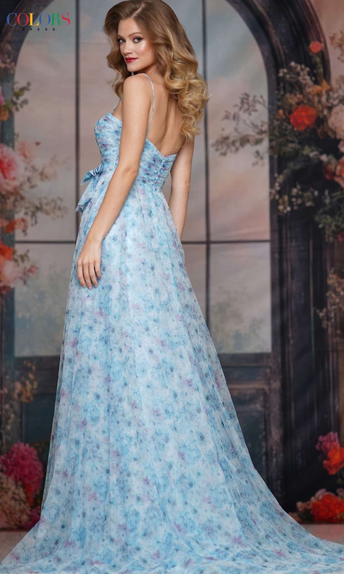 A woman wearing the Strappy Long Print Prom Dress: Colors Dress 3812 stands before a window backdrop decorated with flowers and greenery.